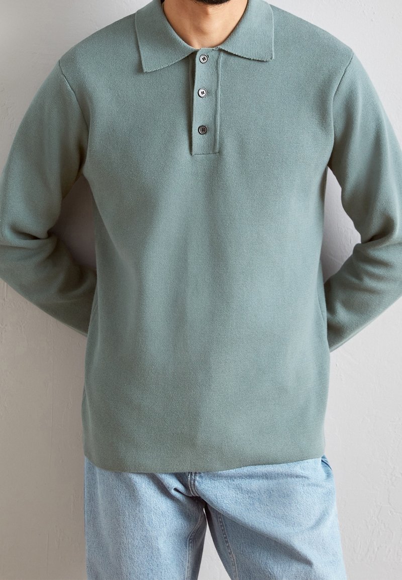 Pull-over vert clair avec un col à revers, une patte à trois boutons et des manches longues. Confectionné en tissu texturé, porté sur un jean bleu clair.