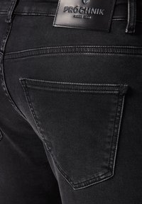 Zbliżenie na czarne dżinsy, tylna kieszeń z przeszyciami i skórzaną naszywką marki z napisem "PRÓCHNIK od 1948".