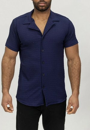 Chemise à manches courtes bleu marine en tissu texturé avec fermeture à boutons, dotée d'un col à revers et d'un ourlet légèrement incurvé.