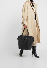 Czarna skórzana torba na ramię z dwoma długimi uchwytami, teksturowaną powierzchnią i metalowym zapięciem, trzymana przez osobę w beżowym trenczu.