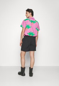 Camisa de manga curta rosa com padrão de rã, calções pretos e botas de borracha pretas. Vista traseira mostrando o design colorido e o ajuste da camisa.