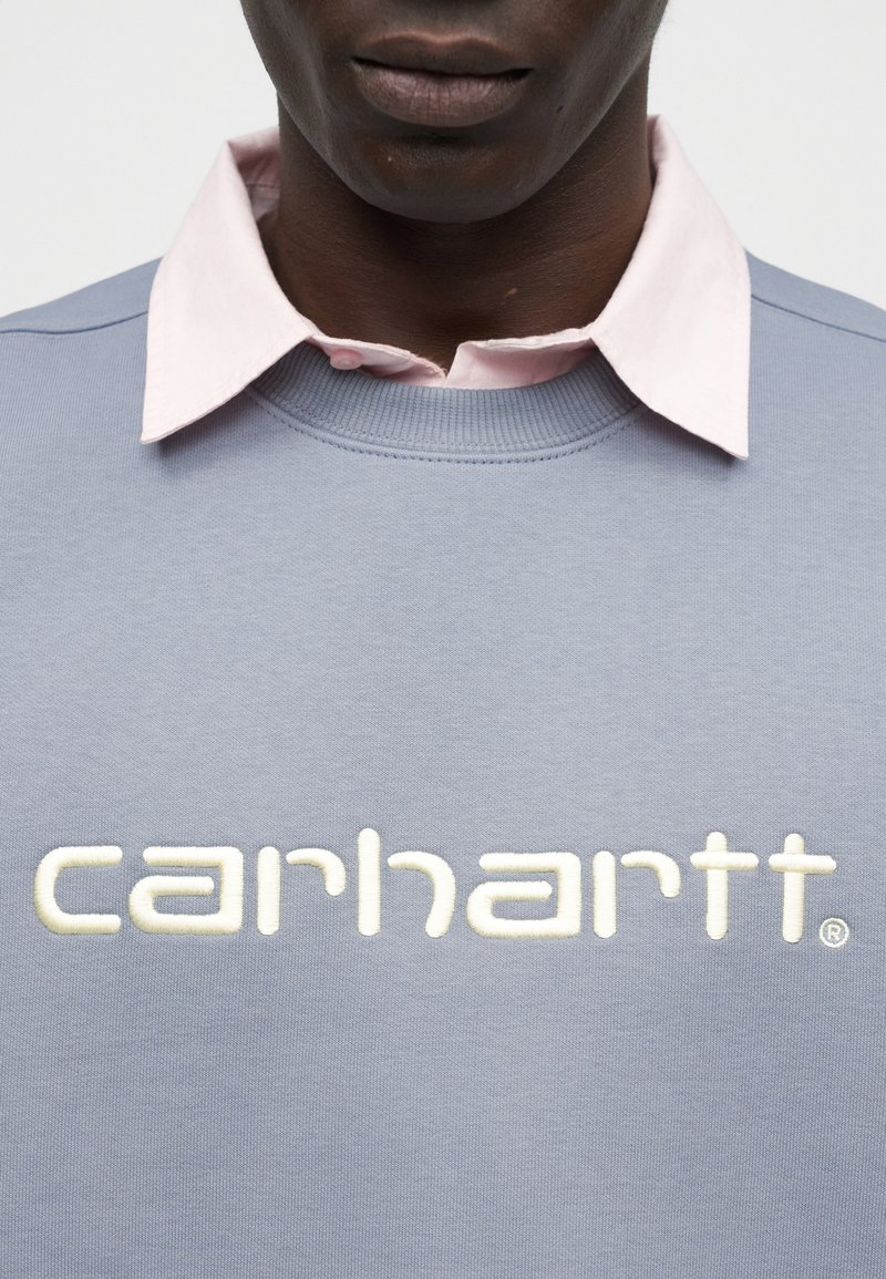 Person trägt einen hellvioletten Pullover mit hervorgehobenem weißem „Carhartt“-Logo und ein blassrosa Hemd mit Kragen darunter.