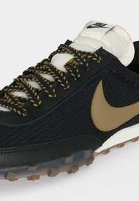 Μαύρο αθλητικό παπούτσι Nike με υφή στην επάνω επιφάνεια, λογότυπο Swoosh σε ταμπά χρώμα, λευκή επένδυση στη γλώσσα και μαύρες και ταμπά πλεκτές κορδόνια σε διαφανές πέλμα με λάστιχα.