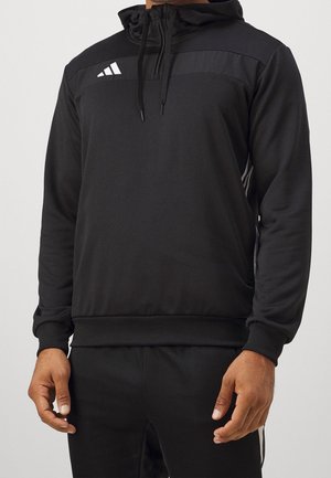 Mann trägt schwarzen Adidas-Hoodie mit weißem Logo auf der Brust und passende schwarze Hose mit weißen Seitenstreifen, steht vor einfarbigem Hintergrund.