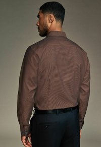 Cămașă lung�ă cu mâneci, de culoare maron cu un model, cu guler tip button-down, asortată cu pantaloni strânși negri și o curea neagră, arătând un material texturat.