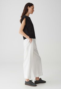 Zwarte mouwloze top met geprofileerde schouderaccenten, gecombineerd met een losse witte broek en donkergrijze sneakers. Minimalistisch ontwerp.