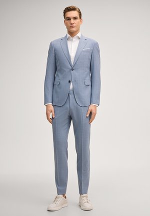 Giovane uomo in piedi che indossa un completo azzurro chiaro, camicia bianca e sneaker bianche su uno sfondo grigio chiaro uniforme.