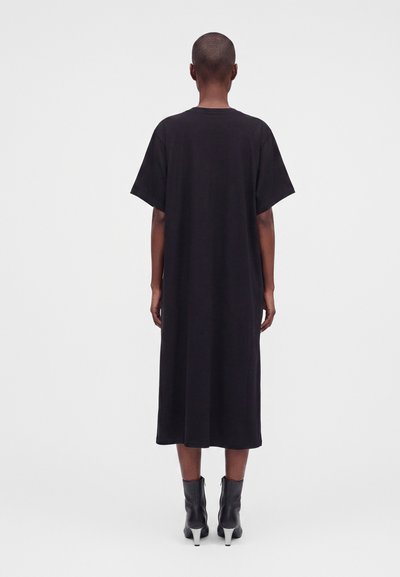Personne debout de dos portant une robe midi noire ample à manches courtes et des bottines noires à talons blancs.