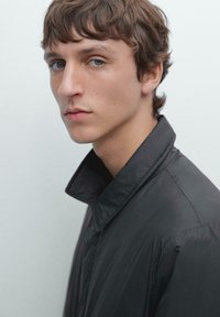 Dunkelgraue Jacke mit hohem Kragen, aus einem glatten, leichten Material; verfügt über ein sauberes, minimalistisches Design. Nahaufnahme eines Menschen.