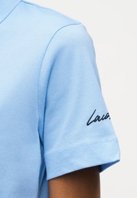 Κοντινή λήψη ενός ανοιχτού μπλε κοντομάνικου πουκάμισου στο μπράτσο ενός ατόμου με μαύρο κείμενο σε πλάγια γραφή τυπωμένο κοντά στο τελείωμα του μανικιού.