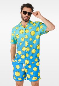 Chemise à manches courtes et shorts assortis en tissu bleu avec imprimé de visages souriants jaunes. Comprend un cordon de serrage blanc sur les shorts.