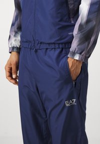 Navy track suit featuring a zip-up jacket with patterned sleeves and elastic waistband. Includes zippered pockets and EA7 logo detail.