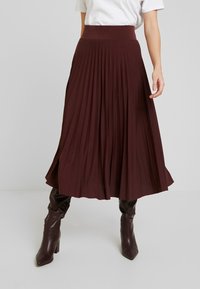 Burgundy veckad kjol med ett brett elastiskt midjeband, tillverkad av ett mjukt, flödande material, som når till mitten av vaden, parat med bruna stövlar som går till knäna.