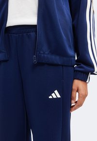 Άτομο που φοράει σκούρο μπλε παντελόνι Adidas και αντίστοιχο μπουφάν με λευκές ρίγες και λογότυπο, στέκεται με χαλαρό χέρι.