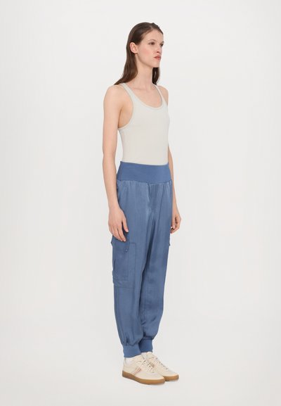 Lichtgrijze tanktop gecombineerd met losse blauwe broek met een elastische tailleband en twee zijzakken, aangevuld met beige sneakers.