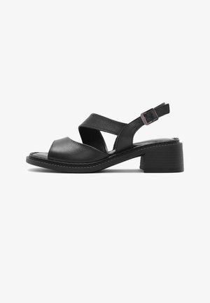 Zwarte leren open teen sandaal met gekruiste bandjes, verstelbare gesp aan de hielriem en lage blokhak op een witte achtergrond.