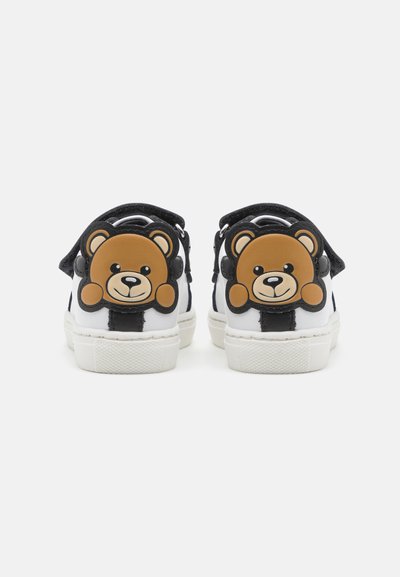 Sneakers bianchi con accenti neri, caratterizzati da un design di orso bruno sul tallone. Materiale liscio e suola testurizzata. Design casual.