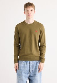 Pull en maille vert olive avec un col rond, des poignets et un ourlet côtelés, arborant un petit logo rouge sur la poitrine gauche. Porté par-dessus un jean bleu clair.