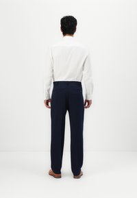 Cămașă albă cu mâneci lungi și pantaloni bleumarin. Pantalonii au buzunare pe spate cu nasturi. Pantofi din piele maro vizibili.