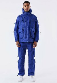 Blauwe weerbestendige jas en broek met meerdere zakken en verstelbare kenmerken, gedragen met witte sneakers. Glad textuur, praktische ontwerpen.