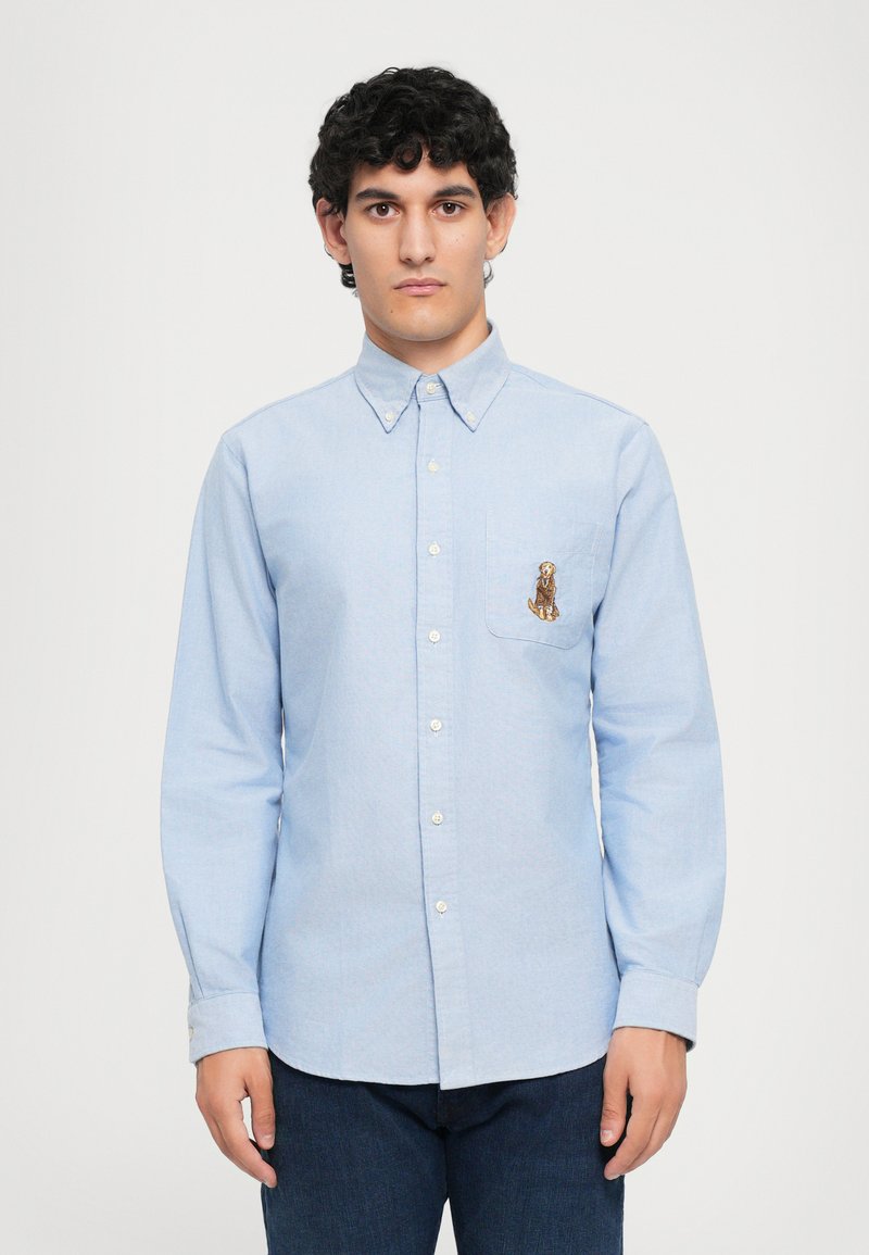 Cămașă bleumarin deschis cu mânecă lungă, cu un buzunar ce prezintă un design brodat cu un câine, fabricată dintr-un material asemănător bumbacului, cu manșete cu nasturi.
