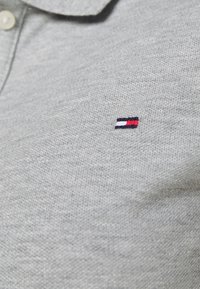 Šedé polo tričko z pleteniny s klasickým límcem. Má malé vyšívané logo v námořnické modré, bílé a červené barvě na hrudi.