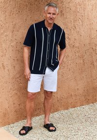 Camisa de manga corta navy con rayas verticales blancas, combinada con pantalones cortos blancos y sandalias negras. Fondo texturizado en tonos cálidos.