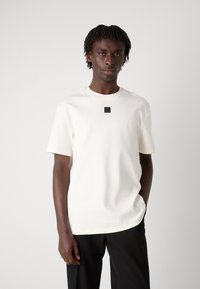 Weißes kurzärmliges T-Shirt aus glattem Stoff, mit einem kleinen schwarzen Logo auf der Brust, kombiniert mit schwarzen Hosen.
