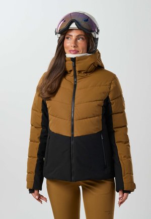 Femme portant une veste de ski marron et noire, un pantalon assorti, un casque blanc et des lunettes de ski réfléchissantes, regardant de côté sur un fond gris.