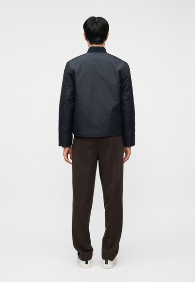 Άνδρας που φοράει μπλε σκούρο μπουφάν, σκούρο καφέ παντελόνι και λευκά αθλητικά παπούτσια με πράσινες και κόκκινες ρίγες, στέκεται μπροστά σε απλό λευκό φόντο.