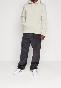 Muž v světlé béžové mikině Adidas, tmavě šedých manšestrových kalhotách a bílých teniskách s černými pruhy, stojí na jednobarevném bílém pozadí.