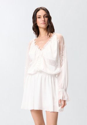 Femme portant un chemisier blanc semi-transparent avec des manches en dentelle et une jupe à volants, se tenant devant un fond clair uni.