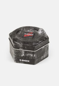 Hexagonal black tin container with a textured surface, featuring a red and white G-Shock logo on the top and a silver metal clasp.
