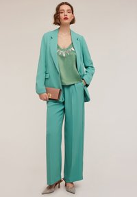 Turkos kavaj och vida byxor i kombination med en grön satinblus med en halsringning prydd med silverpaljetter, samt en texturerad clutch i roséguld.