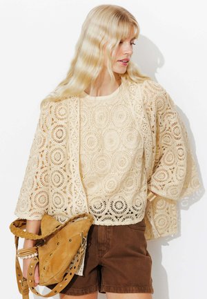 Femme blonde portant un haut beige en crochet et une veste assortie, un short marron, tenant un sac en daim beige avec une bandoulière tressée et portant des bracelets.