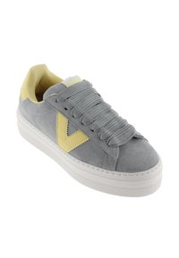 Sneaker in suede grigio con suola in gomma bianca, caratterizzata da lacci grigi e un accento a forma di V giallo sul lato. Design semplice con punta rotonda.