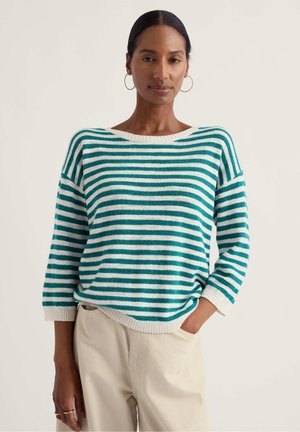 Femeie purtând un pulover cu dungi albastre și albe cu mâneci trei sferturi și pantaloni bej, stând cu o mână în buzunar.