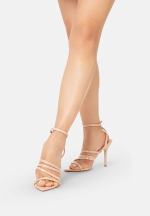 Piernas cruzadas con sandalias nude de tacón alto y tiras con cierre en el tobillo sobre un fondo liso y claro.