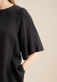 Blusa nera in lino con maniche corte a sbuffo e scollatura rotonda. Il tessuto ha una leggera texture ed è dotata di una discreta tasca laterale.
