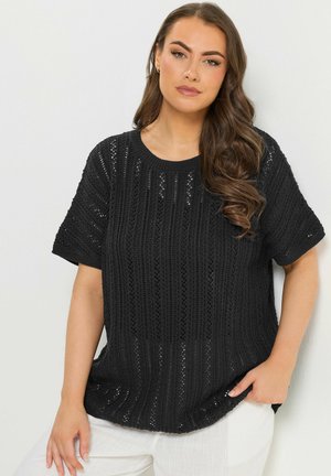 Kobieta z długimi falującymi włosami, ubrana w czarny dziany top z krótkim rękawem i białe spodnie, stojąca na jasnym jednolitym tle.