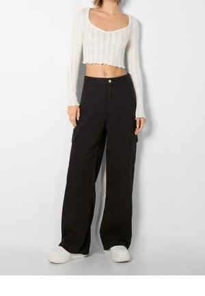 Top corto blanco con textura acanalada y escote redondeado, combinado con pantalones cargo negros holgados que tienen bolsillos laterales y cierre de botón. Zapatillas blancas.