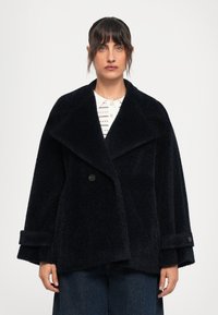 Casaco oversized azul-marinho, semelhante a pelo, com lapelas largas, fecho de um único botão e punhos dobrados, combinado com uma camisa branca de colarinho por baixo.