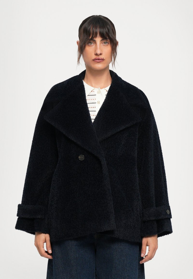 Casaco oversized azul-marinho, semelhante a pelo, com lapelas largas, fecho de um único botão e punhos dobrados, combinado com uma camisa branca de colarinho por baixo.