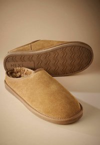 Tan suede slippers with a soft interior lining, rounded toe, and a textured rubber sole featuring a wavy pattern. Brand name visible on the sole.