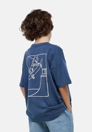 Personne aux cheveux bouclés portant un t-shirt bleu avec un dessin blanc représentant un skateboarder réalisant une figure sur une rampe.