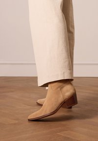 Stivaletti beige in suede con punta affusolata e tacco a blocco marrone texturizzato, abbinati a pantaloni larghi color beige chiaro su un pavimento di legno.