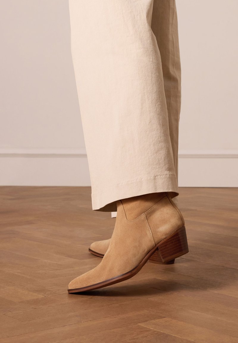 Stivaletti beige in suede con punta affusolata e tacco a blocco marrone texturizzato, abbinati a pantaloni larghi color beige chiaro su un pavimento di legno.