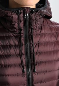Chaqueta acolchada color burdeos con un collar alto, que presenta una cremallera central y detalles de cordón. La superficie tiene una textura suave y brillante con un patrón acolchado.