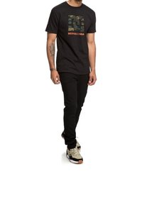 Black short-sleeve t-shirt featuring a camo graphic and orange text, paired with black pants and multicolored sneakers.