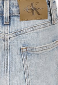 Jeans in denim azzurro chiaro con un'etichetta in pelle beige che mostra "Calvin Klein Jeans" in rilievo, dotati di tasche standard e cuciture.